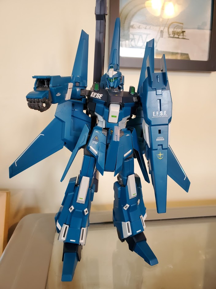 Photo of an MG ReZEL Commander Gundam model kit. It is holding a shield up with its left arm and is holding its right arm straight forward to point its arm rockets.